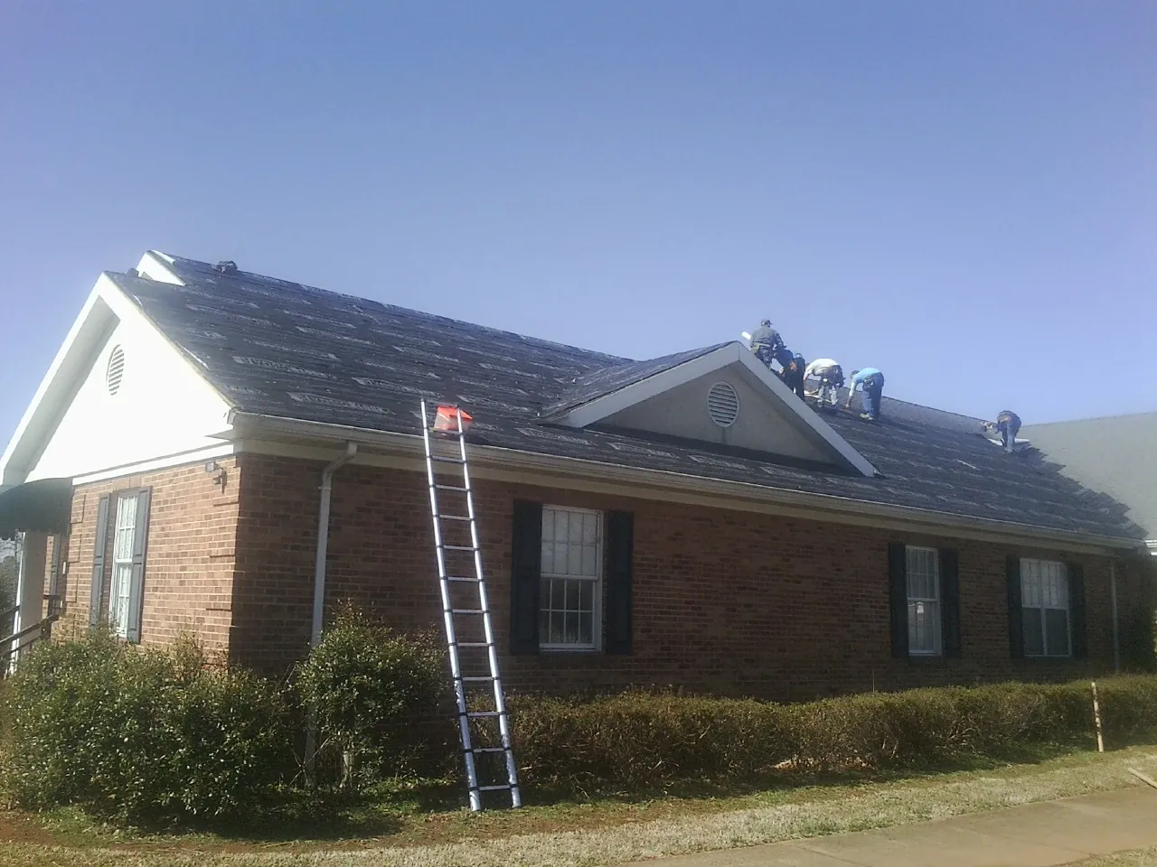 Crew tearing shingles off a brick ranch roof with a ladder set — Emergency Roof Repair work by our crew in Saint Georges