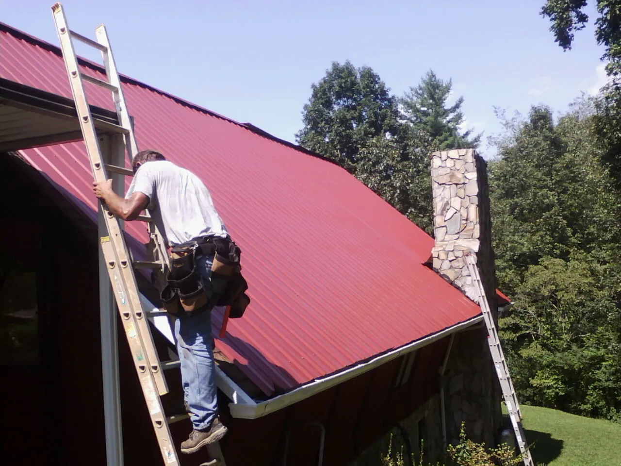 Roofer on a ladder beside a red metal roof and stone chimney — Soffit & Fascia Repair by Roofing Brothers in Saint Georges